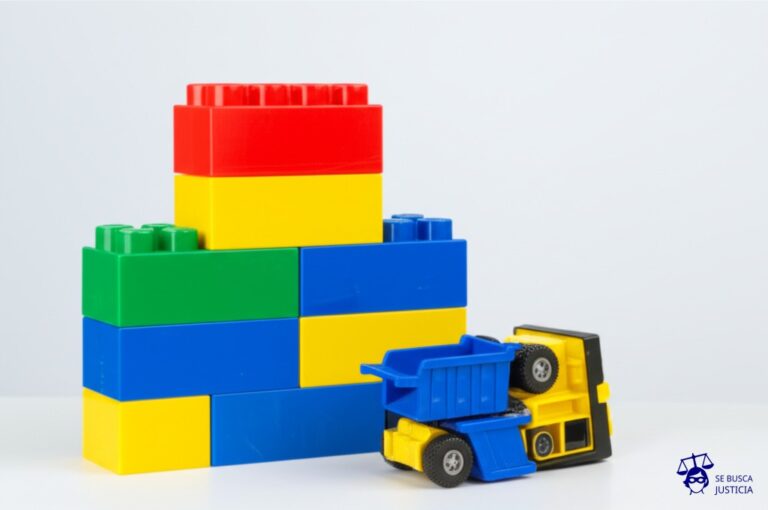 Un camión de juguete, aplastado bajo una pila de bloques de construcción desproporcionadamente grandes. Representa: Accidente en transporte de carga por sobrepeso