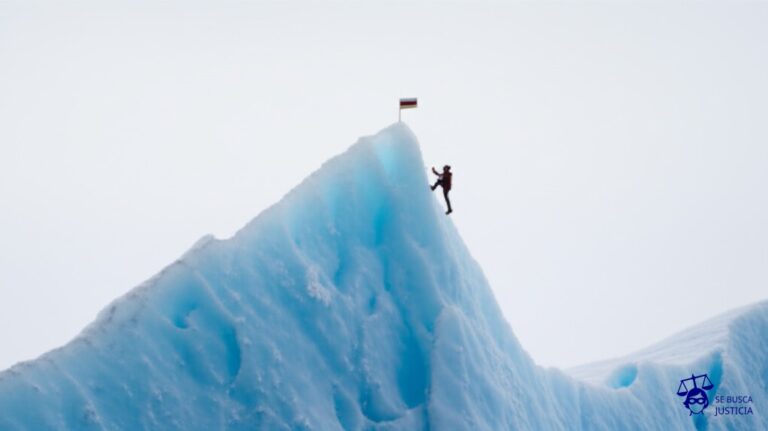 Un gran iceberg con grietas, en el que hay una figura humana diminuta intentando escalar con dificultad, con una pequeña banderita clavada en la cima. Representa: Marina Abramović enfrentó demandas por lesiones durante algunas de sus performances extremas