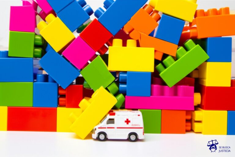 Una gran pila de bloques de construcción infantiles (de colores brillantes), desmoronándose y cayendo, bloqueando el camino a un pequeño coche de juguete (ambulancia). Representa: Obstáculos al acceso humanitario en territorios en conflicto