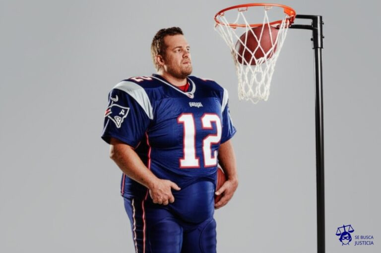 Un hombre robusto, con una camiseta de fútbol americano a medio inflar, intentando desesperadamente meter una pelota desinflada en una canasta de baloncesto. Representa: Sanción a Tom Brady por el "Deflategate" en la NFL